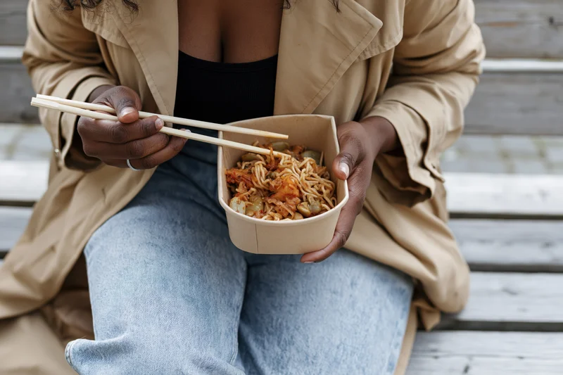 Woman eating noodles from a takeout bowl with chopsticks while sitting on a wooden bench