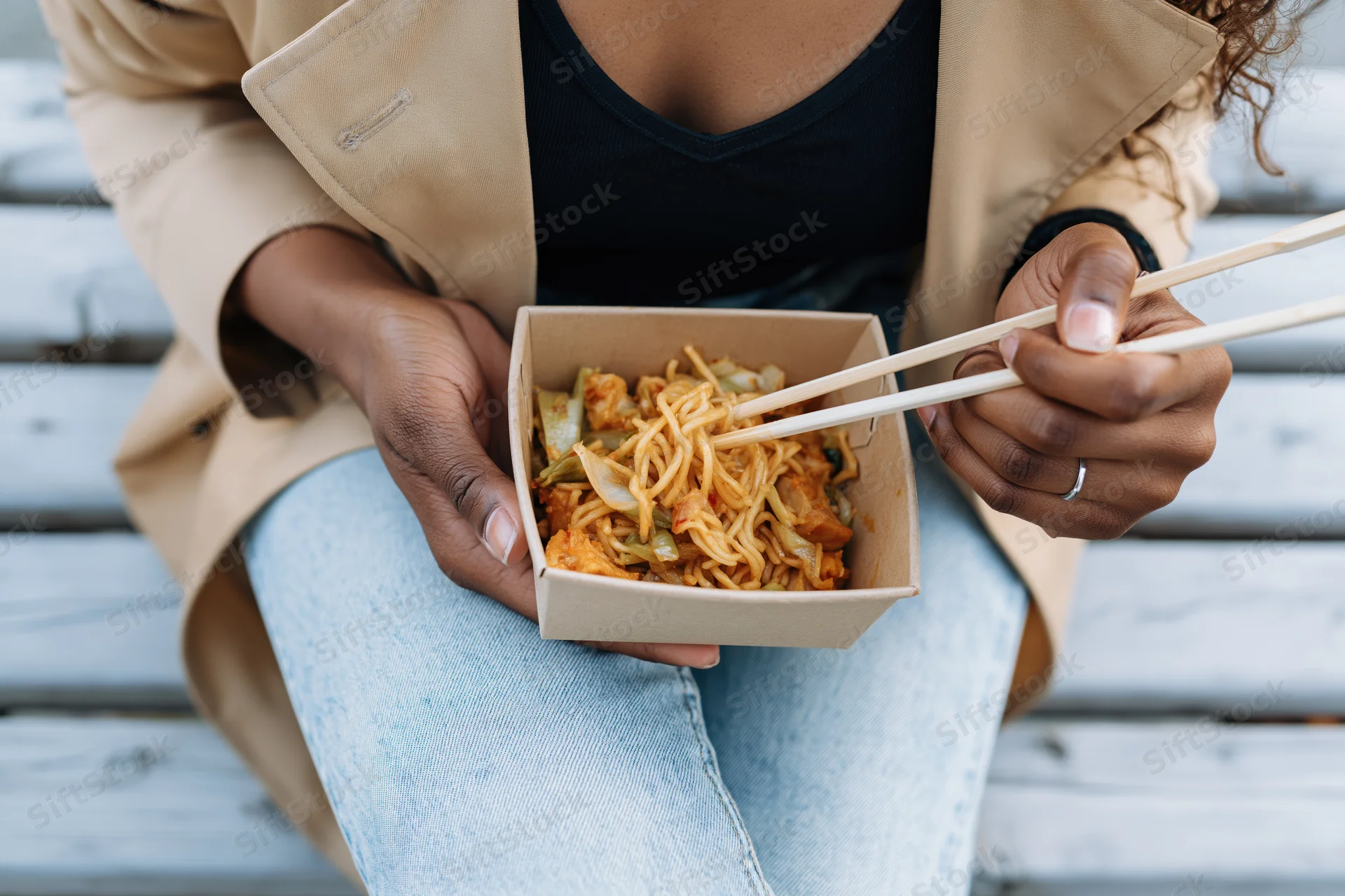 Woman eating noodles from a takeout box with chopsticks on a wooden bench