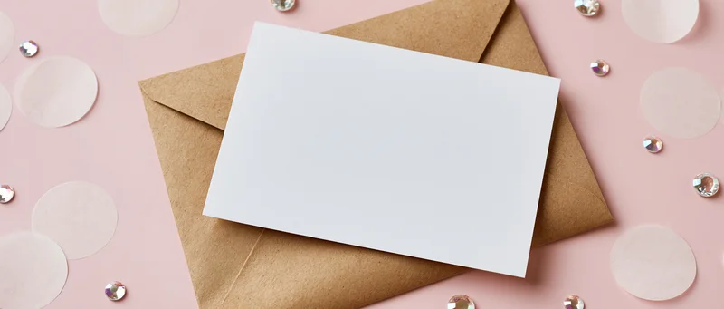 Blank white greeting card on kraft envelope with rhinestones and confetti on pink surface