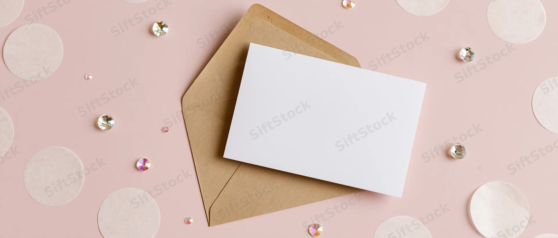 Blank white greeting card on kraft envelope with rhinestones and polka dots on pink surface