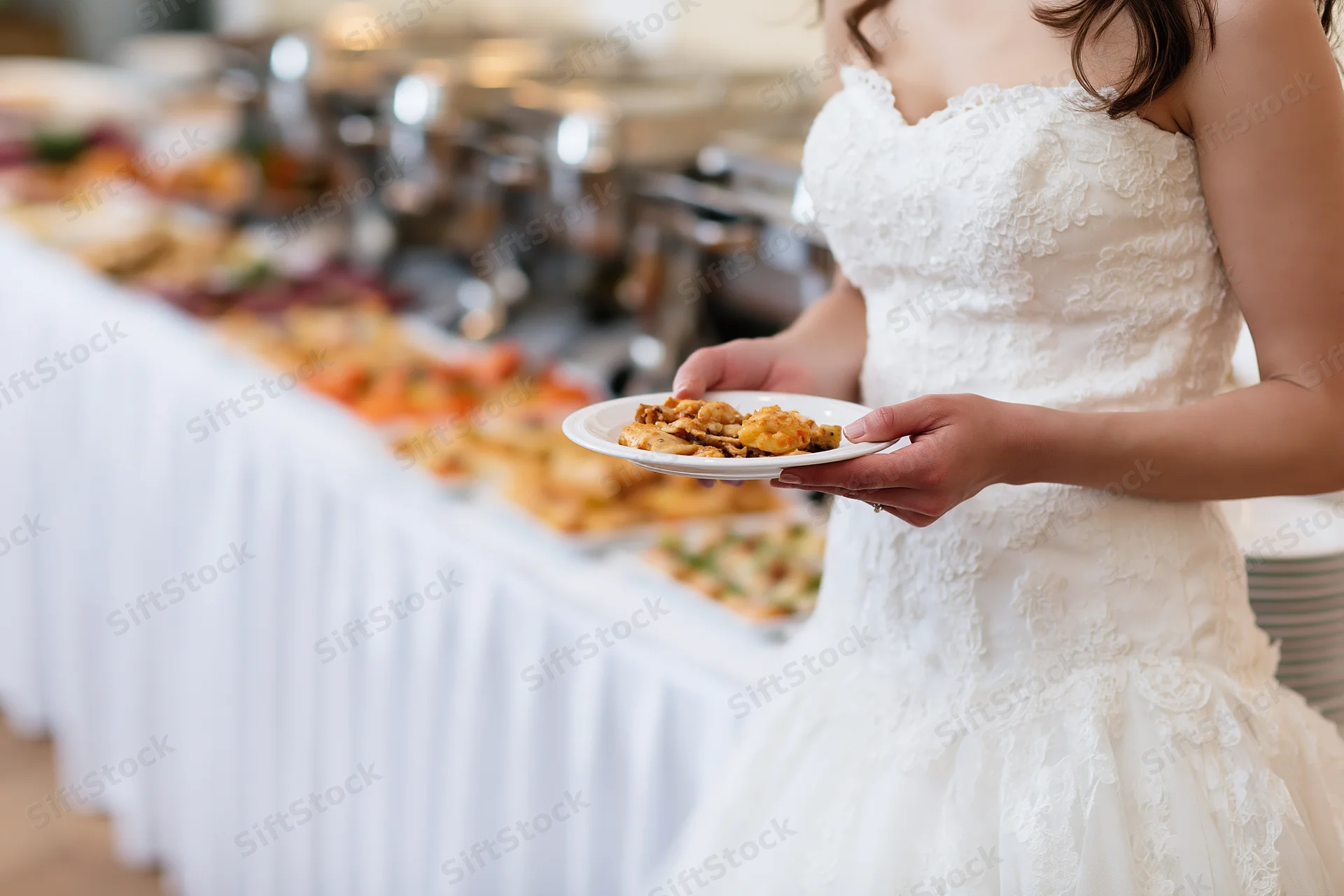 Bride in white lace gown holding plate of food at wedding reception buffet