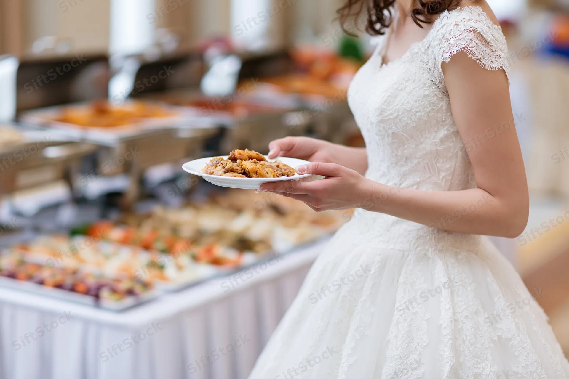 Bride in white lace gown holding plate of food at wedding reception buffet