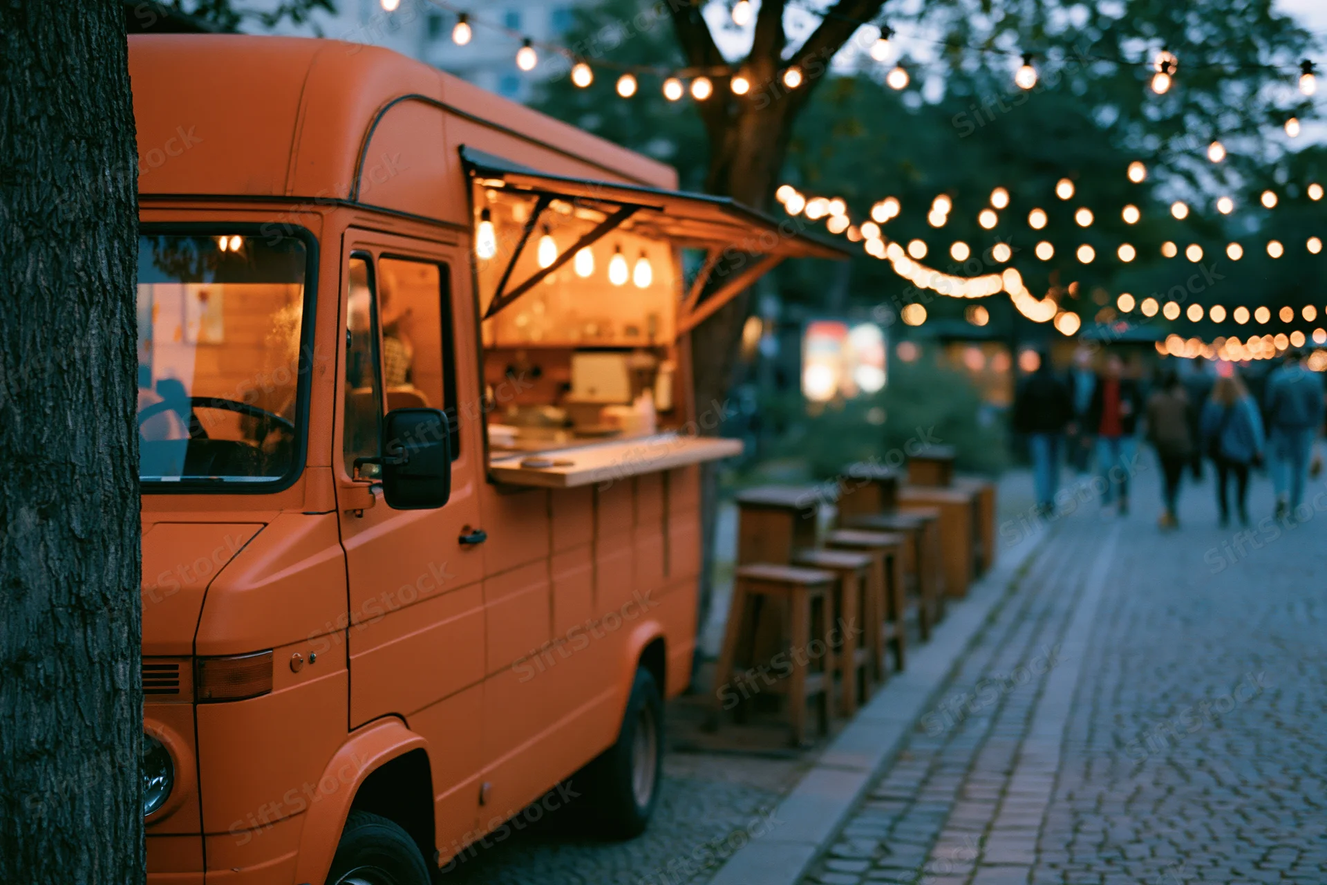 Orange food truck with open awning and string lights along cobblestone street at evening