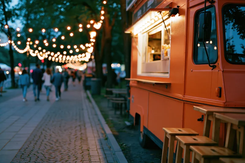 Bright orange food truck parked along cobblestone sidewalk at evening with string lights and pedestrians walking by
