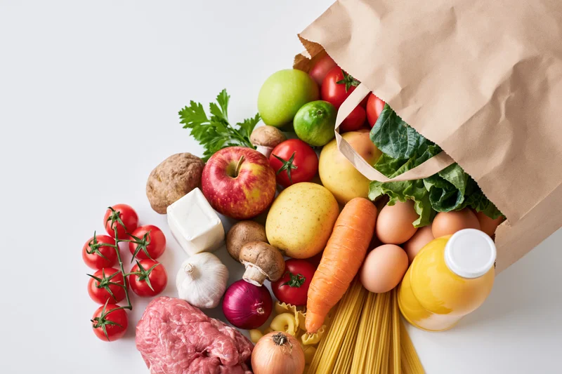 Brown paper grocery bag tipped over spilling fresh produce and groceries onto white surface