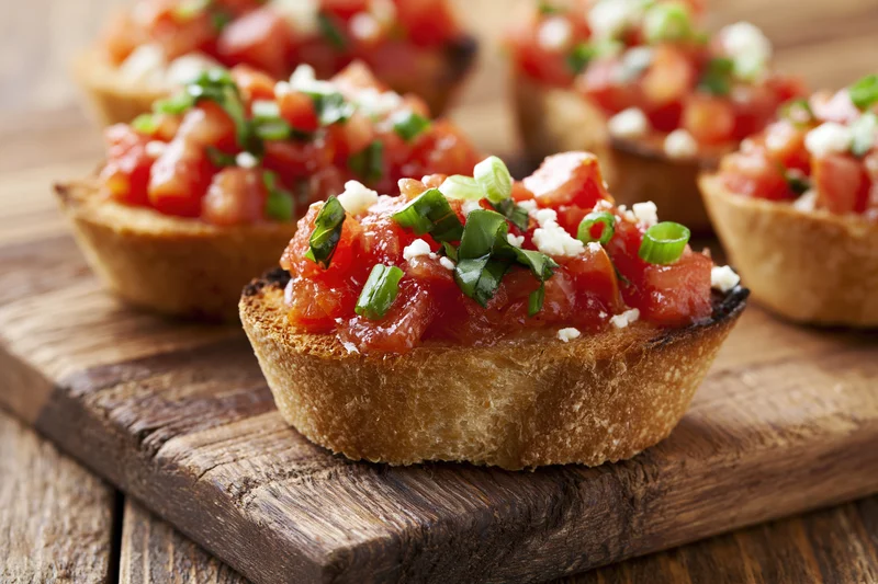 Bruschetta appetizers topped with diced tomatoes, basil, and cheese on a rustic wooden cutting board
