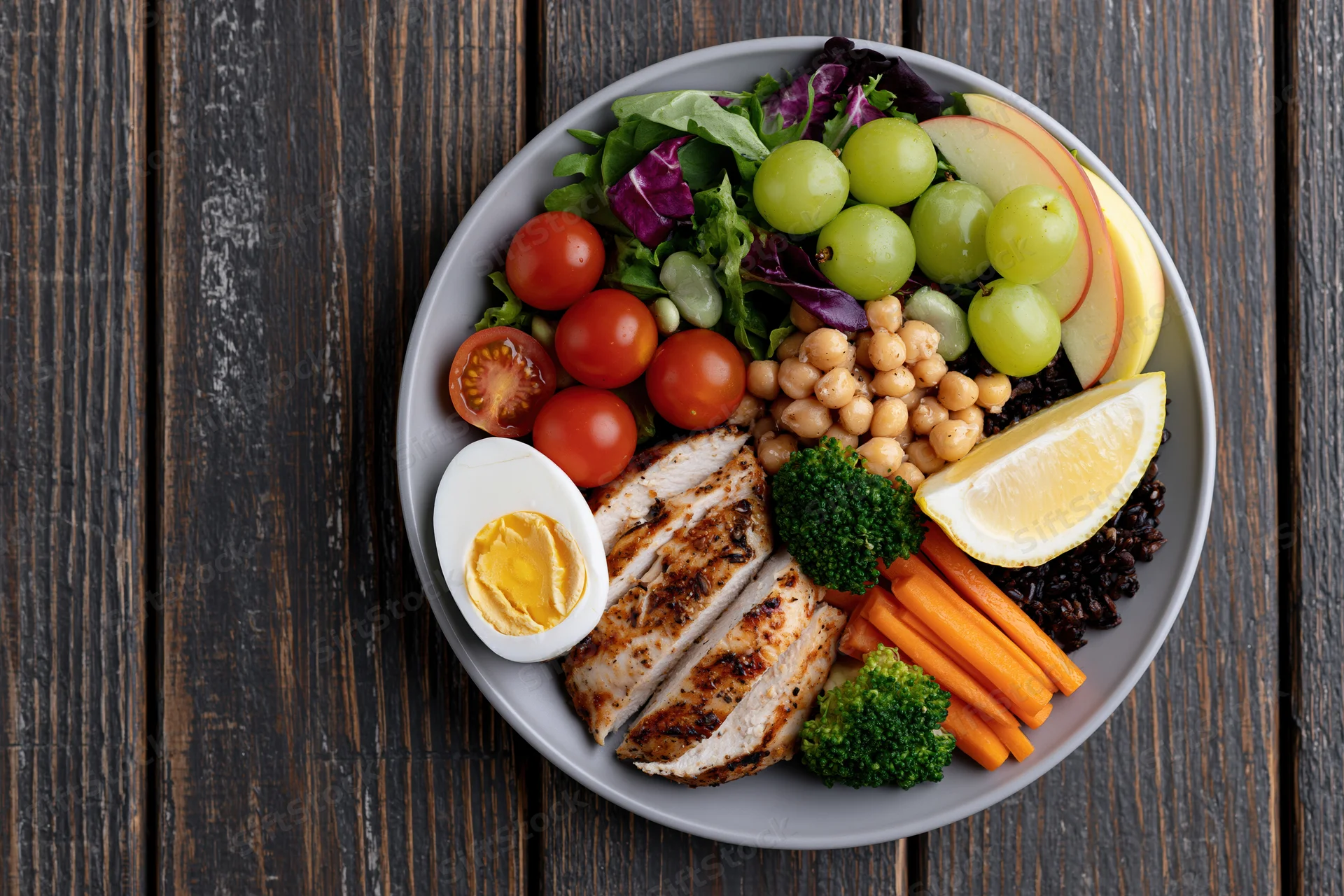 Buddha bowl with grilled chicken, vegetables, fruits and chickpeas on a grey plate atop dark wooden table