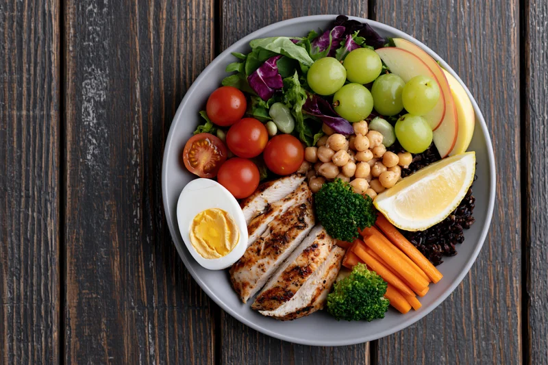 Buddha bowl with grilled chicken, vegetables, fruits and chickpeas on a grey plate atop dark wooden table