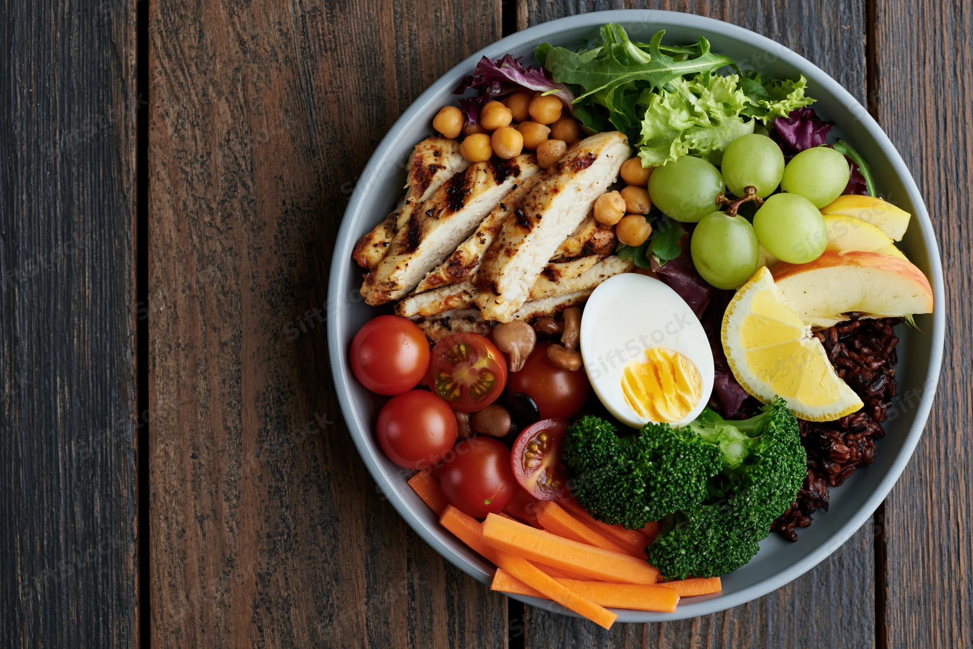 Buddha bowl with grilled chicken, vegetables, fruit, and egg on dark wooden table, overhead view