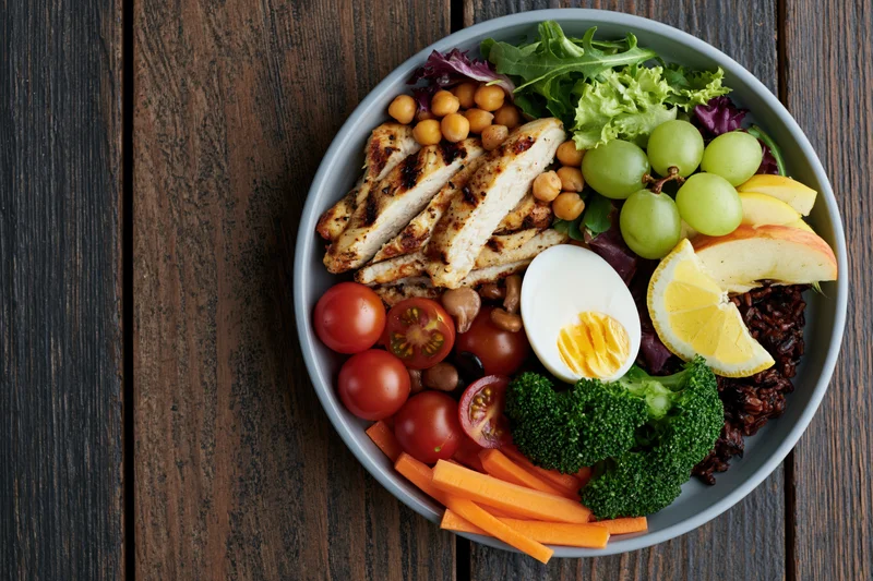 Buddha bowl with grilled chicken, vegetables, fruit, and egg on dark wooden table, overhead view
