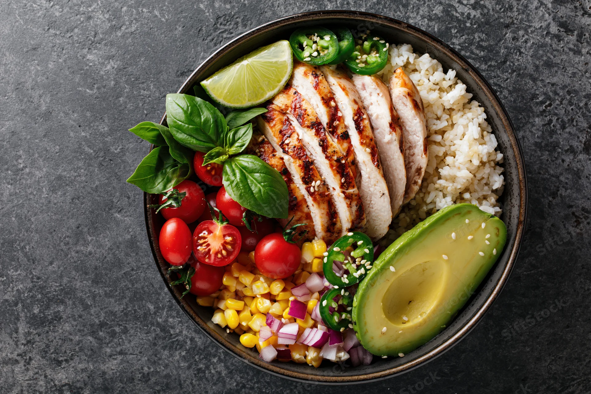 Overhead view of a buddha bowl with grilled chicken, rice, avocado, tomatoes, corn, basil, lime, and jalapeno on a dark textured surface