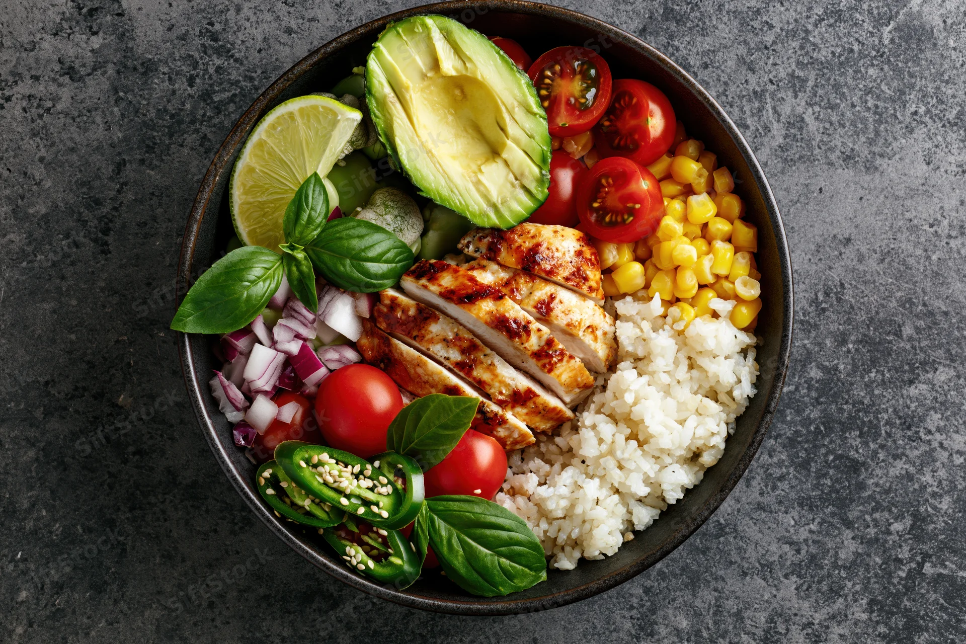 Overhead view of a buddha bowl with grilled chicken, rice, avocado, corn, tomatoes, lime, basil and jalapeno on a dark stone surface