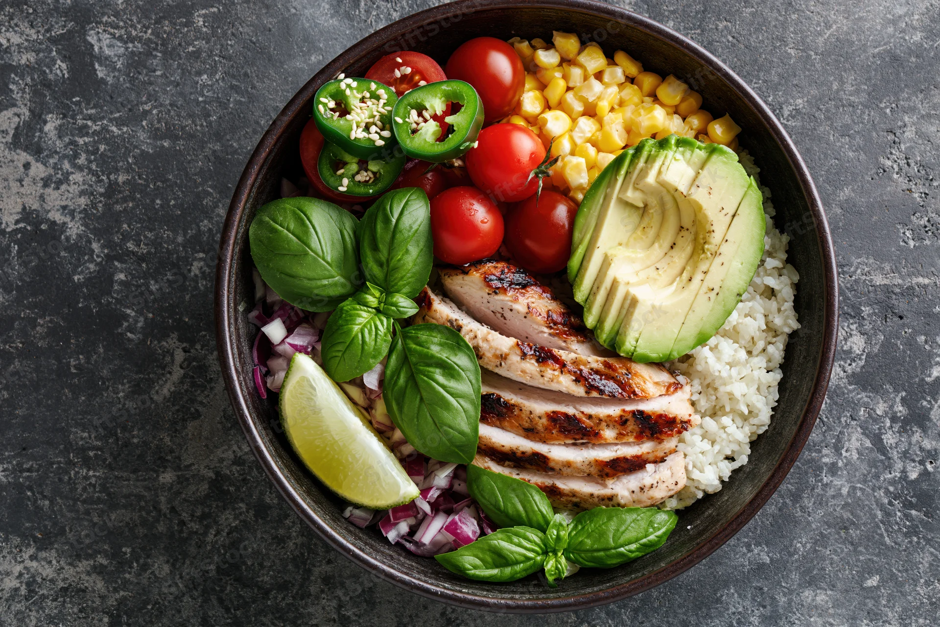 Overhead view of a buddha bowl with grilled chicken, rice, avocado, corn, tomatoes, jalapeno, basil, lime, and red onion on a dark textured surface