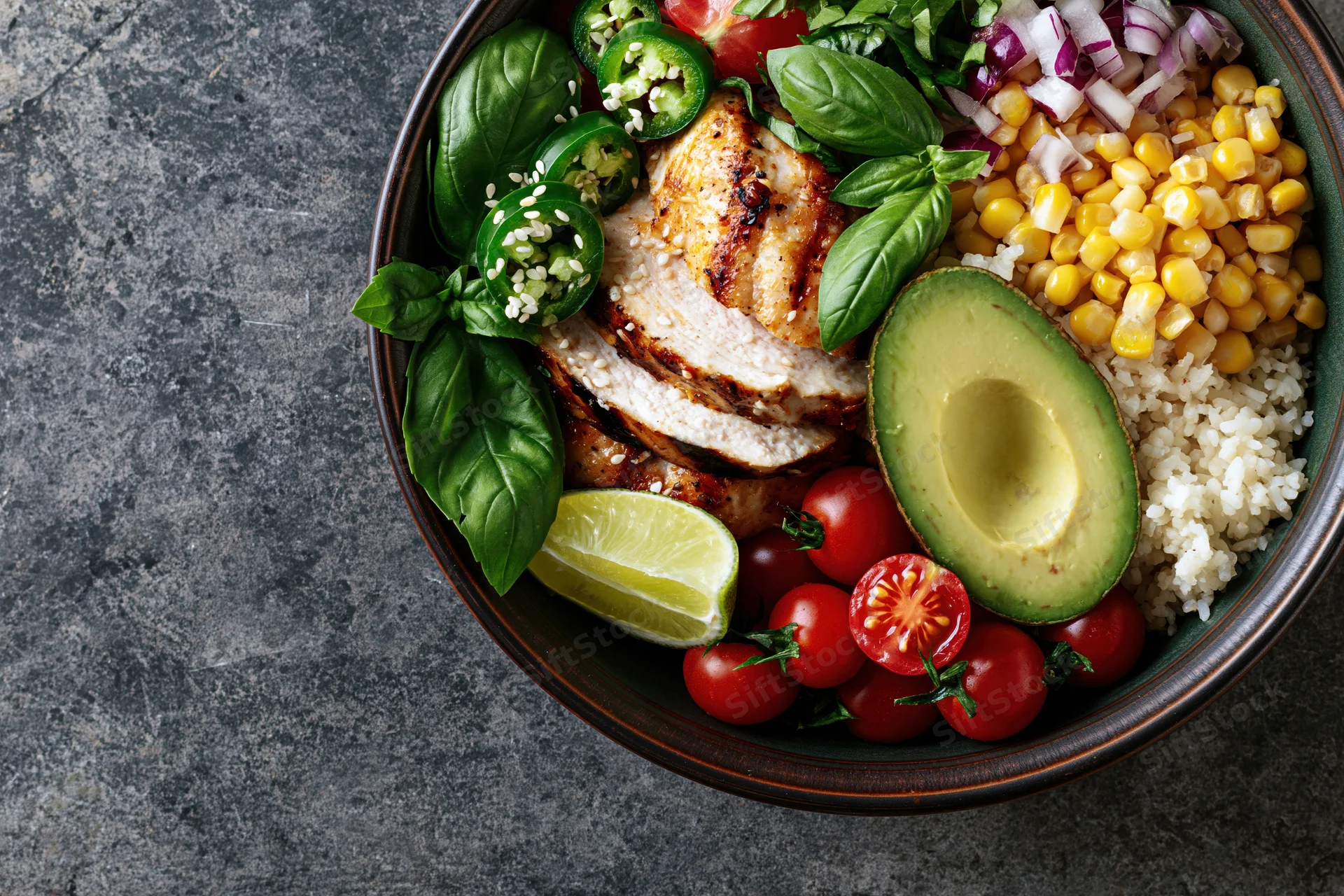 Buddha bowl with grilled chicken, avocado, rice, corn, tomatoes, lime, basil and jalapeno on gray stone surface