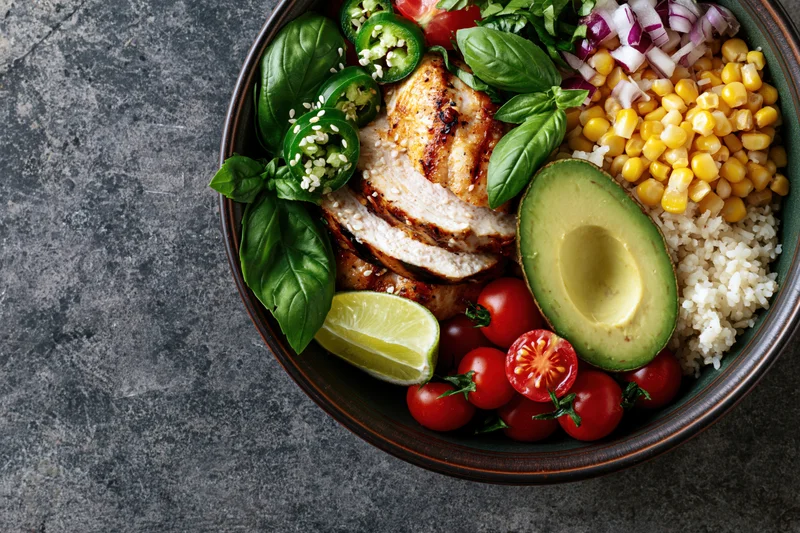 Buddha bowl with grilled chicken, avocado, rice, corn, tomatoes, lime, basil and jalapeno on gray stone surface