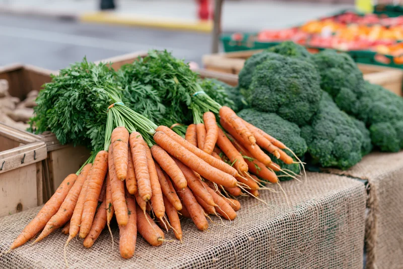 Bunches of fresh carrots with leafy tops and heads of broccoli displayed on burlap at a farm market stall