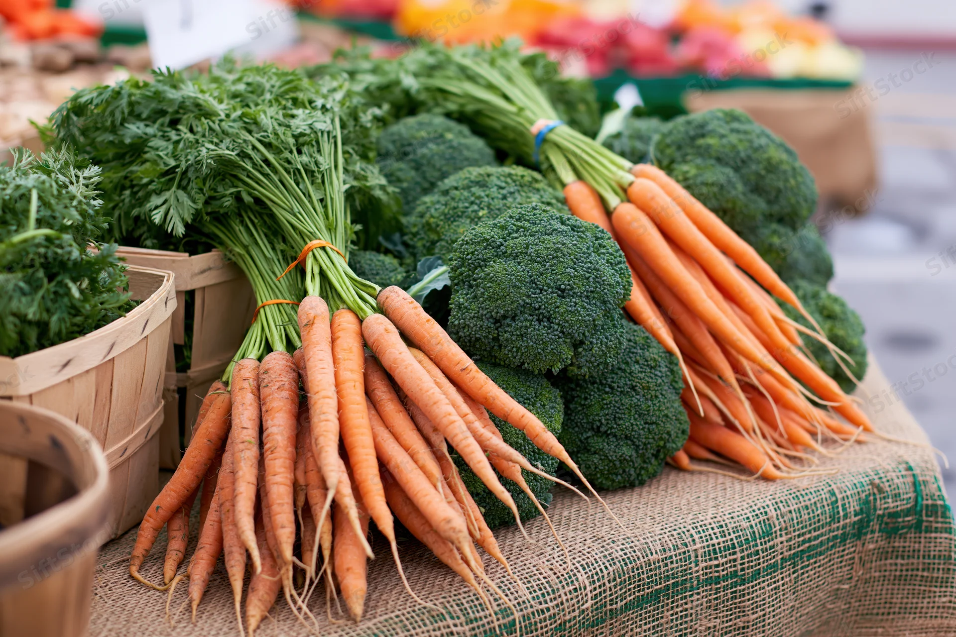 Bunches of fresh carrots with leafy tops and heads of broccoli displayed on a burlap-covered table at a farm market stall