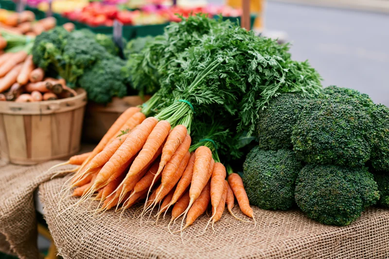 Bunches of fresh carrots with leafy tops and heads of broccoli arranged on a burlap-covered table at an outdoor market stall