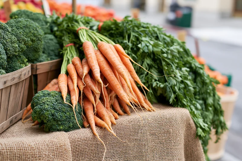 Bunches of fresh carrots with leafy tops and heads of broccoli arranged on a burlap-covered table at a farm market stall