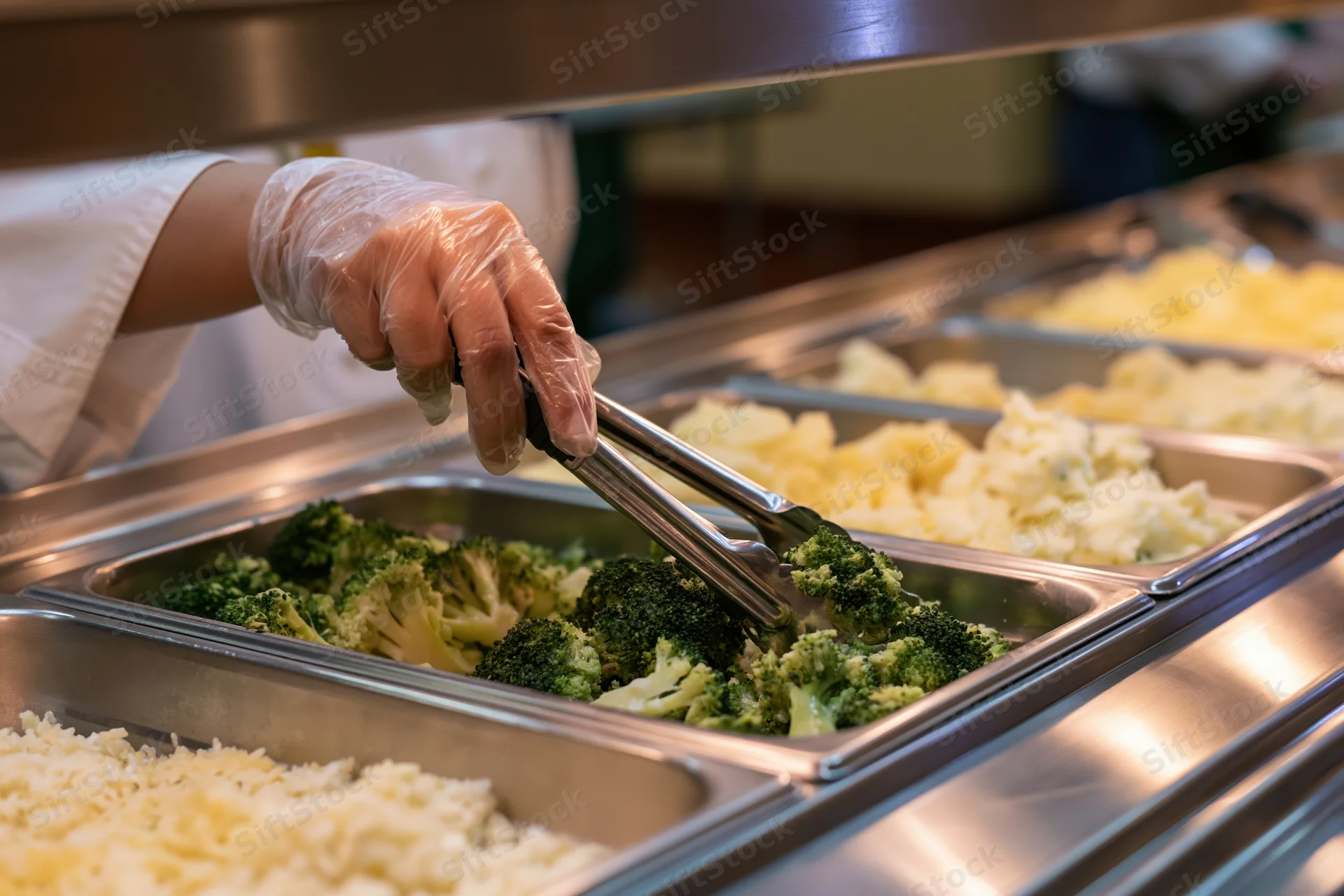 Cafeteria worker using tongs to serve broccoli from a stainless steel buffet line
