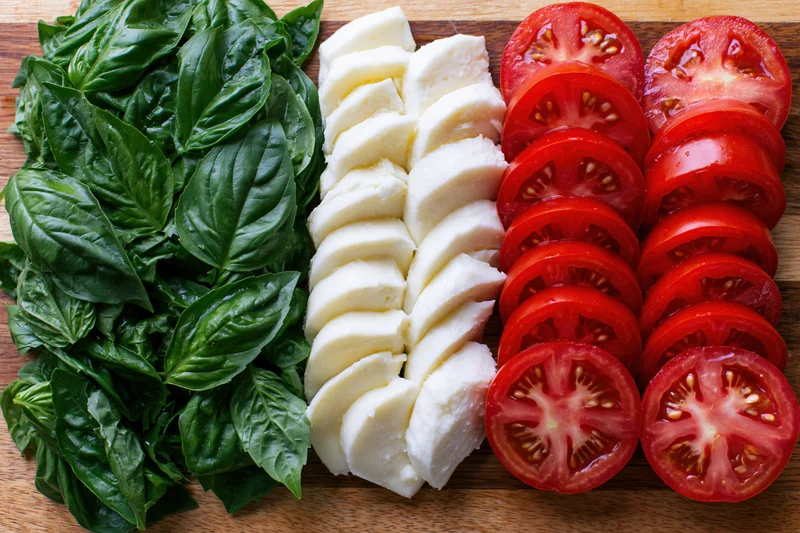 Sliced tomatoes, mozzarella cheese, and fresh basil arranged in parallel rows on a wooden cutting board