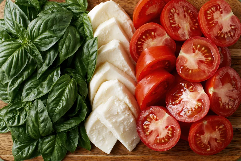Sliced tomatoes, fresh basil leaves and mozzarella cheese arranged in parallel rows on a wooden cutting board