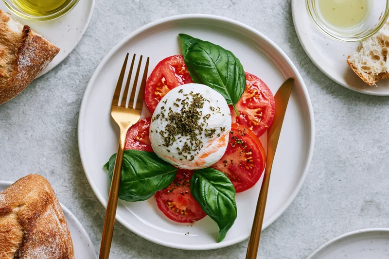 Caprese salad with mozzarella, tomatoes, and basil on a white ceramic plate with gold cutlery, surrounded by bread and olive oil, overhead view