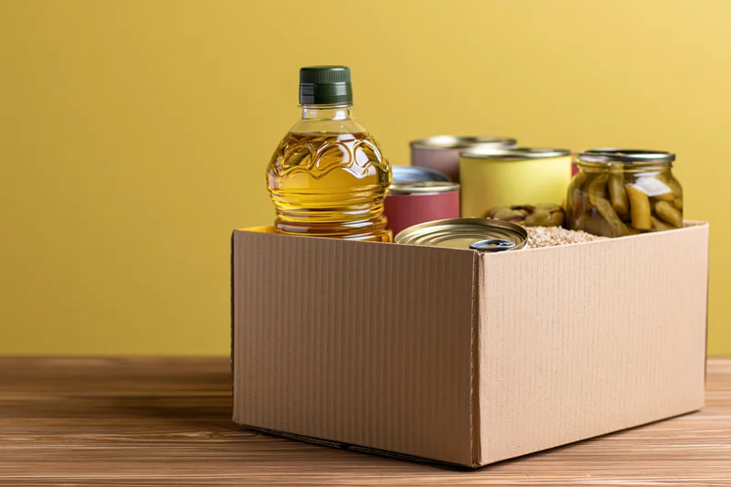 Cardboard box filled with pantry staples including cooking oil and canned goods on wooden table