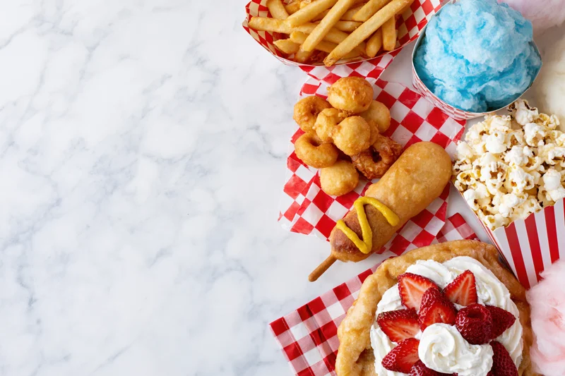 Carnival fair food spread including funnel cake, corn dog, fries, popcorn, and cotton candy arranged along the right edge on a marble surface