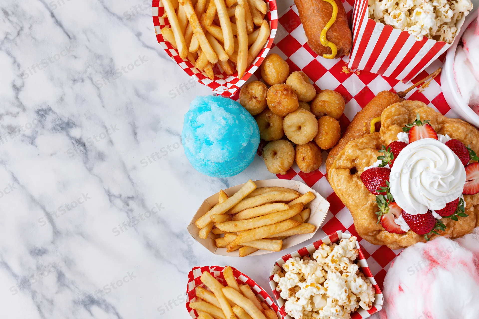 Carnival fair food spread arranged along right edge of marble table with red and white checkered paper baskets and cotton candy, funnel cake, fries, popcorn, and donuts