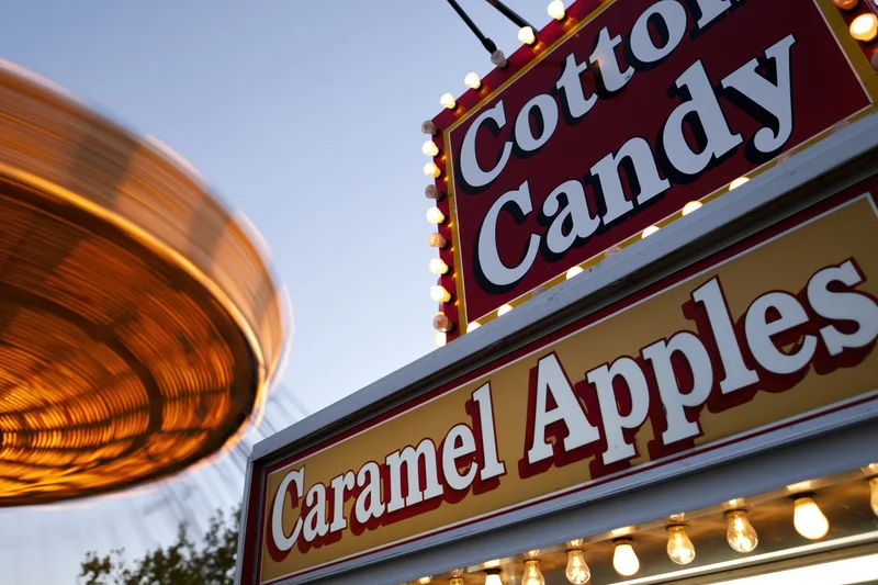 Illuminated carnival food stand signs for cotton candy and caramel apples with a spinning ride in motion against twilight sky