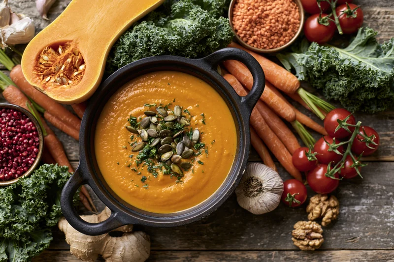 Smooth orange pumpkin soup in a cast iron pot surrounded by fresh vegetables on a wooden table