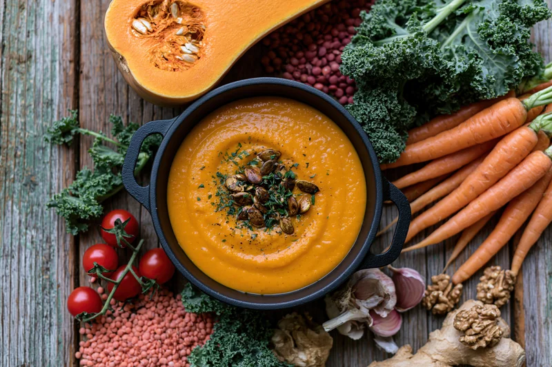 Smooth orange pumpkin soup in a cast iron pot garnished with seeds and herbs surrounded by fresh vegetables on a wooden table
