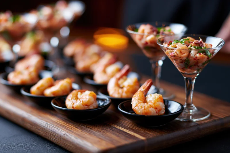 Shrimp appetizers arranged on a dark wooden board with cocktail glasses and small black bowls
