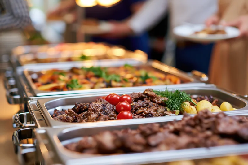 Grilled meat and roasted vegetables on a buffet line with people serving themselves in the background