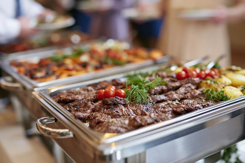 Grilled meat and roasted vegetables served on stainless steel trays at a catering buffet line