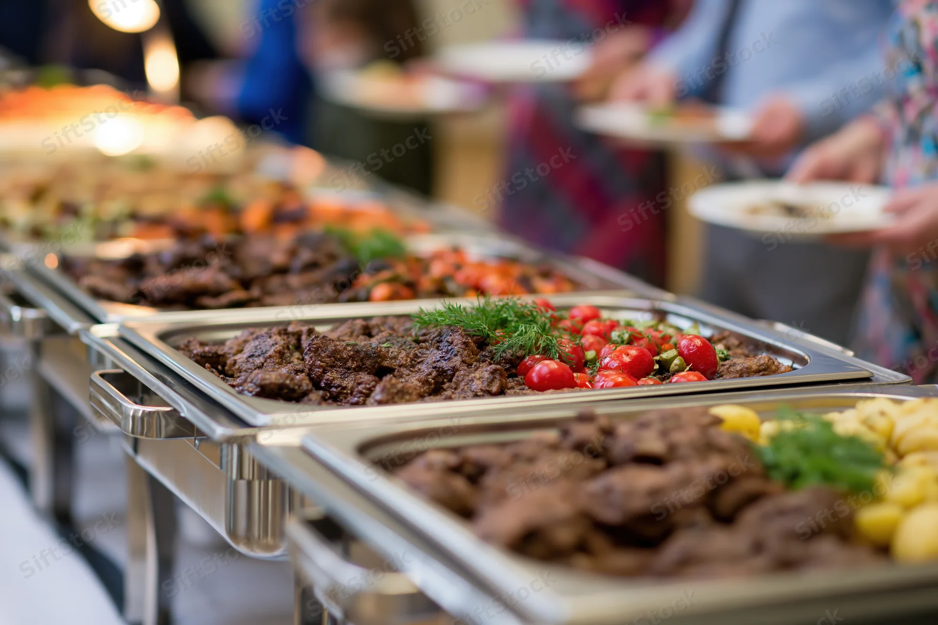 Grilled meat and roasted vegetables served on stainless trays at a buffet line with people holding plates in the background