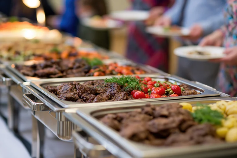 Grilled meat and roasted vegetables served on stainless trays at a buffet line with people holding plates in the background