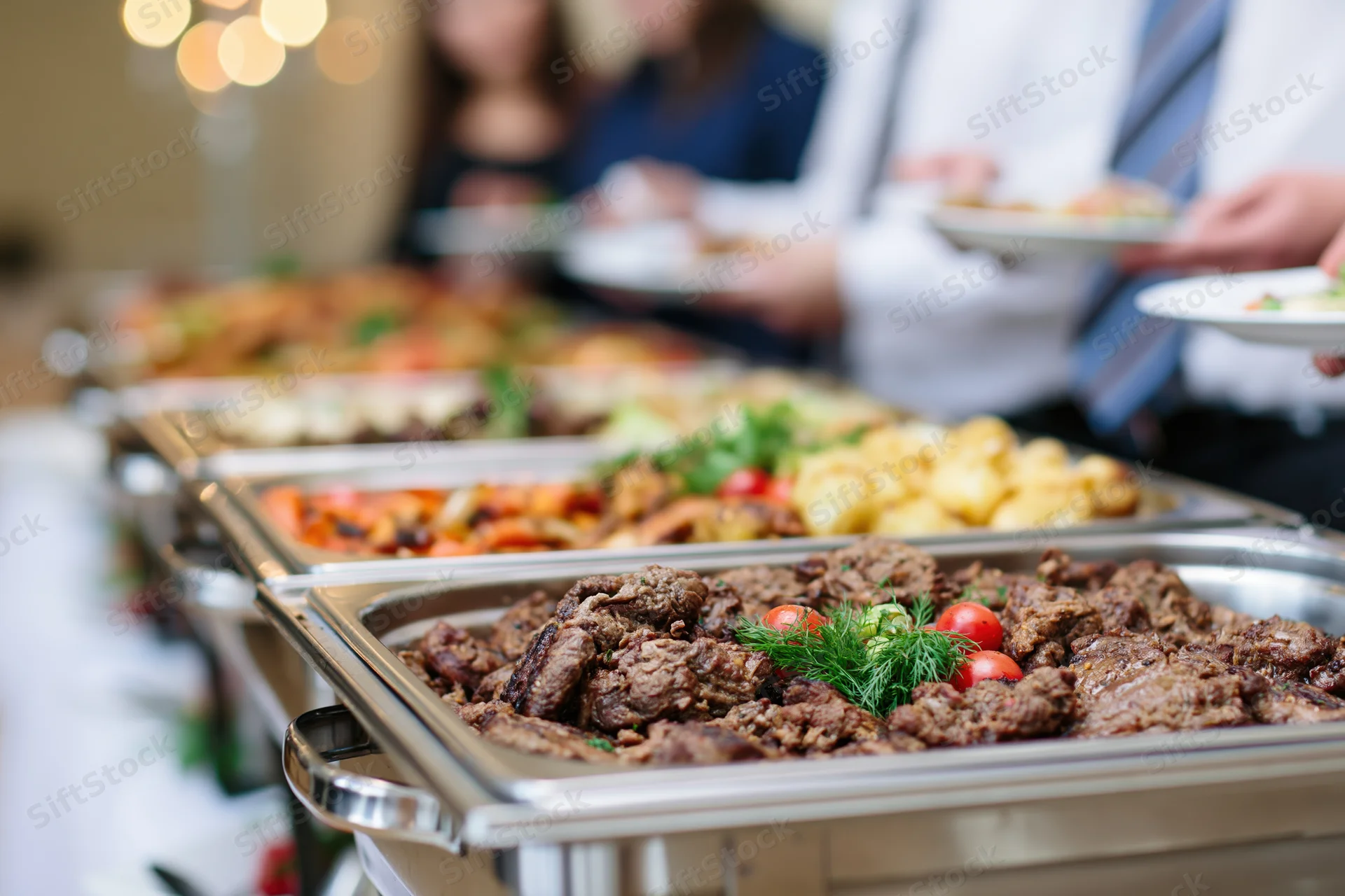 Grilled beef and roasted vegetables served on stainless trays at a catered buffet line with guests in formal attire selecting food