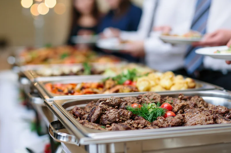 Grilled beef and roasted vegetables served on stainless trays at a catered buffet line with guests in formal attire selecting food