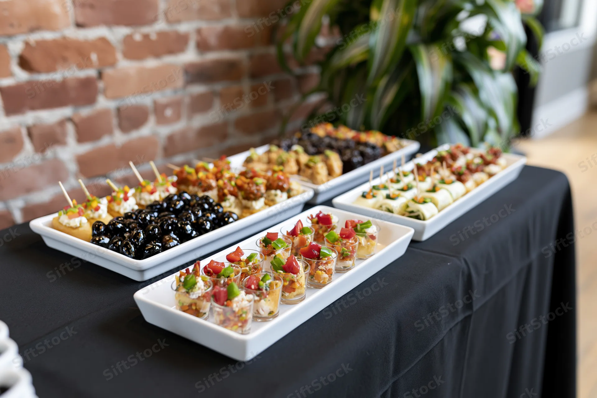 Catering buffet table with appetizers on white platters and glass cups arranged on black tablecloth indoors