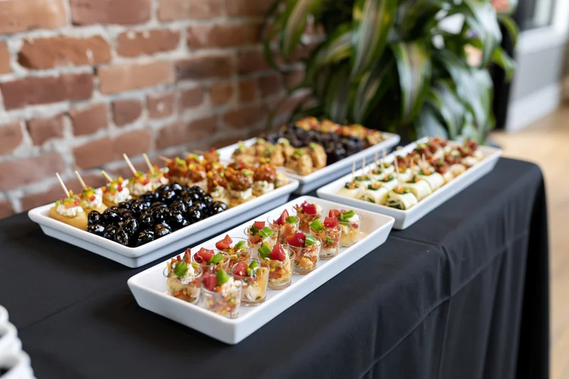 Catering buffet table with appetizers on white platters and glass cups arranged on black tablecloth indoors