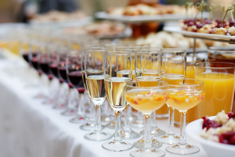 Rows of champagne flutes and glasses with red wine, juice, and cocktails on a buffet table at a reception