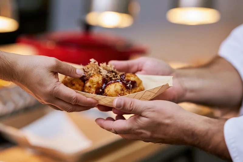 Chef handing a paper boat tray of takoyaki topped with dark sauce and shredded cabbage to a customer in a restaurant kitchen