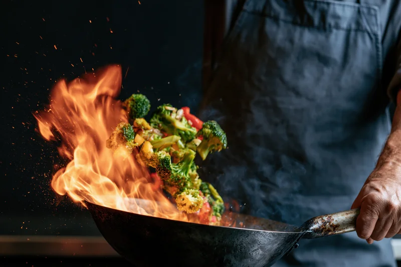 Chef tossing stir-fry vegetables mid-air in a wok with flames and sparks in a dark kitchen