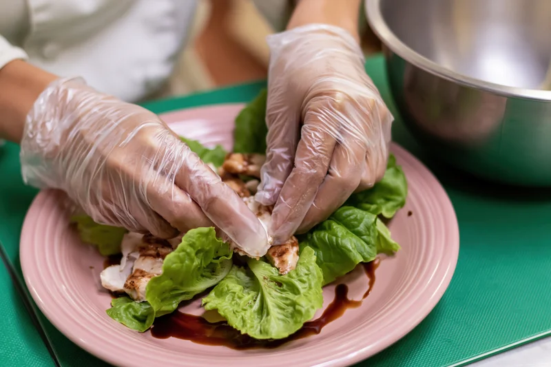 Chef's gloved hands arranging grilled chicken and lettuce on a pink plate over a green cutting board