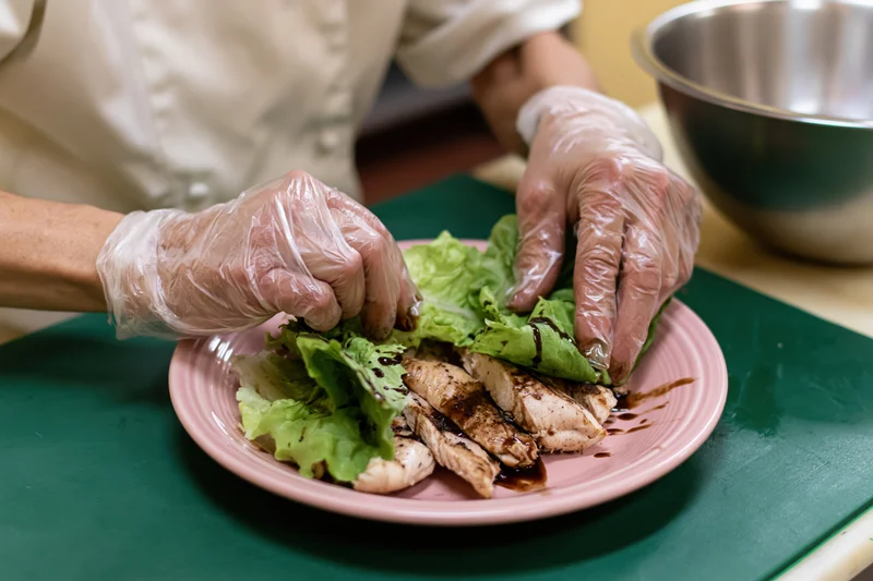 Chef’s gloved hands arranging grilled chicken and lettuce on a pink plate over a green cutting board