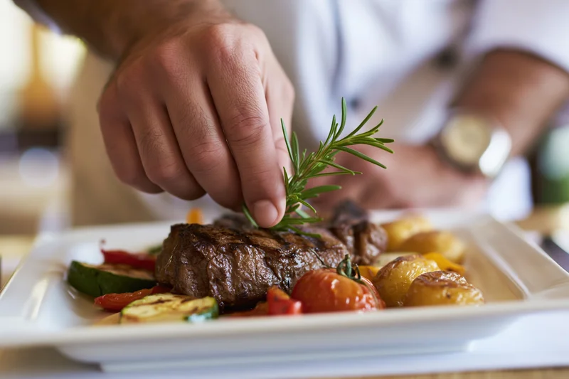 Chef placing a rosemary sprig on a grilled steak with vegetables on a white plate