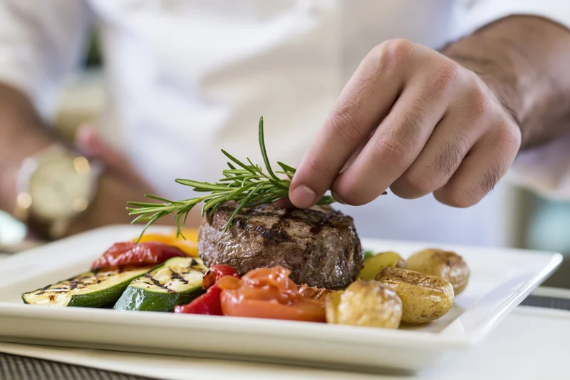Chef placing rosemary garnish on grilled steak with vegetables on white plate