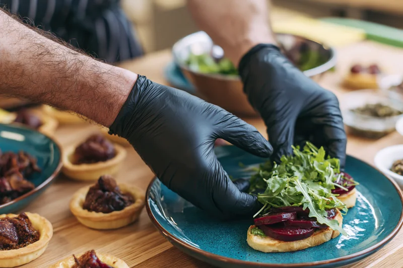 Chef's hands in black nitrile gloves arranging fresh arugula on a beet sandwich on a blue plate at a dining table